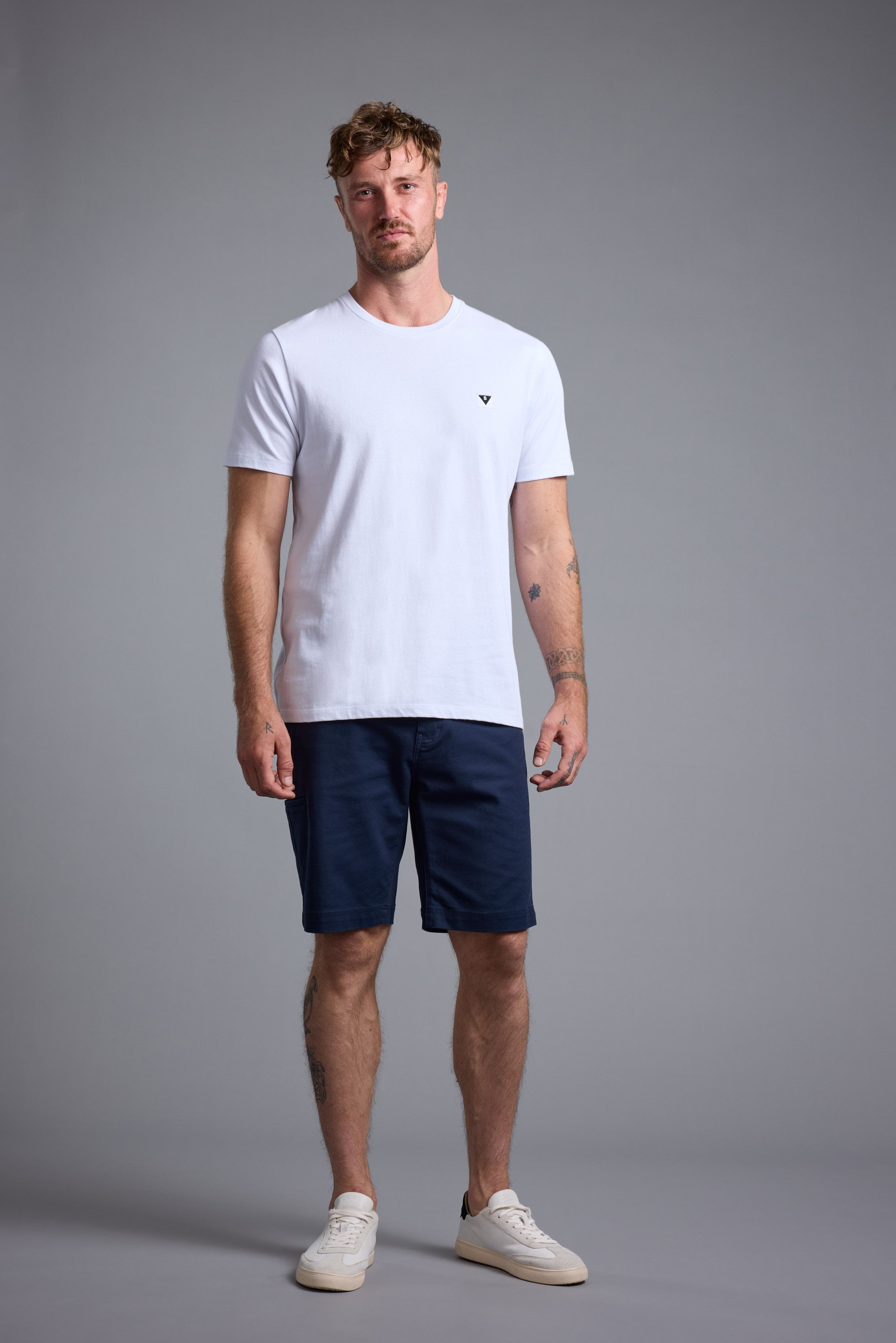 A man wears a white t-shirt, Barkers Anvil 5Pkt Shorts in navy, and white sneakers, standing facing forward against a gray background with a neutral expression and relaxed posture.