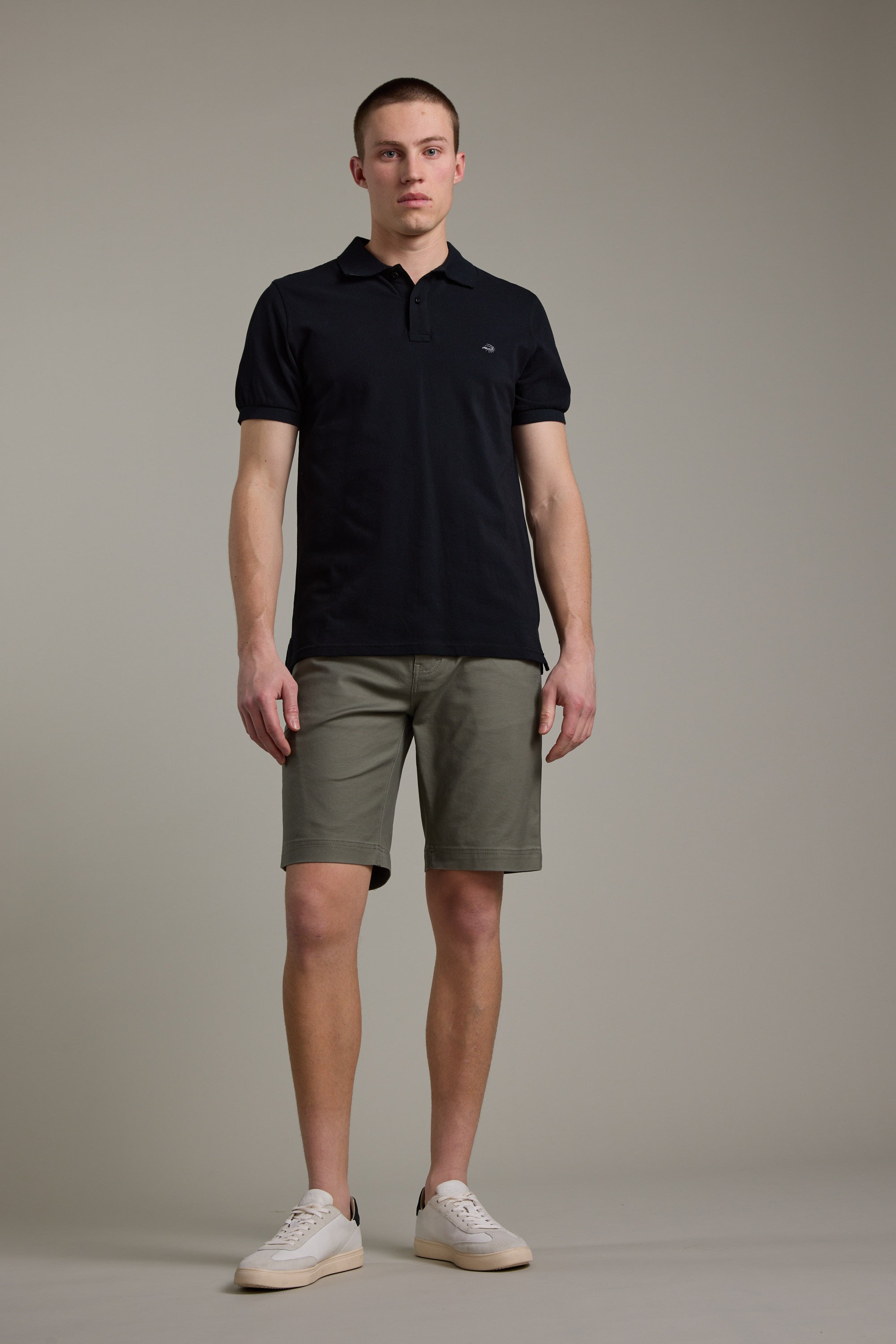 A man stands against a plain background wearing Barkers Anvil 5Pkt Shorts with an elasticated waistband, a black polo shirt, and white sneakers. He faces forward with a neutral expression.