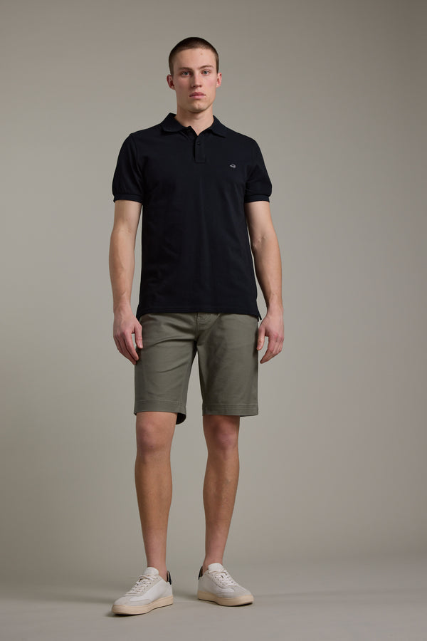 A man stands against a plain background wearing Barkers Anvil 5Pkt Shorts with an elasticated waistband, a black polo shirt, and white sneakers. He faces forward with a neutral expression.