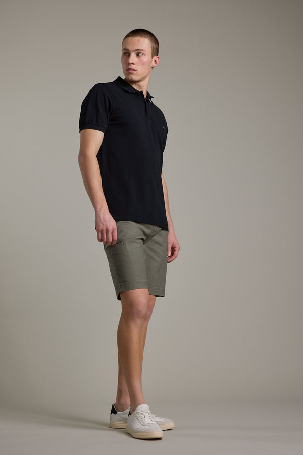 A man stands against a plain background wearing a black polo shirt, Barkers Anvil 5Pkt Shorts with an elasticated waistband, and white sneakers. He looks slightly to the side with one leg extended forward.