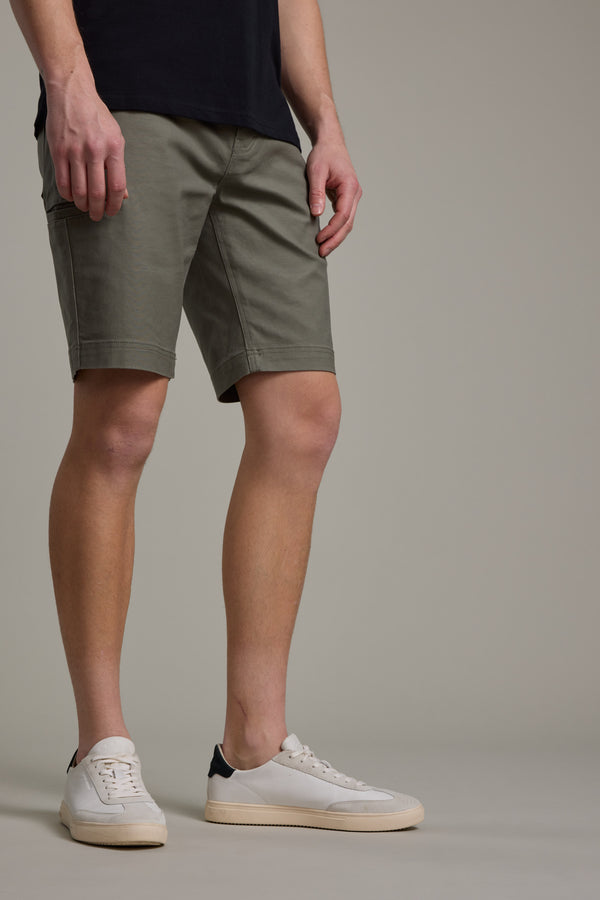 Someone wears the Barkers Anvil 5Pkt Short in olive green with an elasticated waistband, paired with a dark shirt and white sneakers with beige soles, standing against a plain light gray background, cropped below the shoulders.