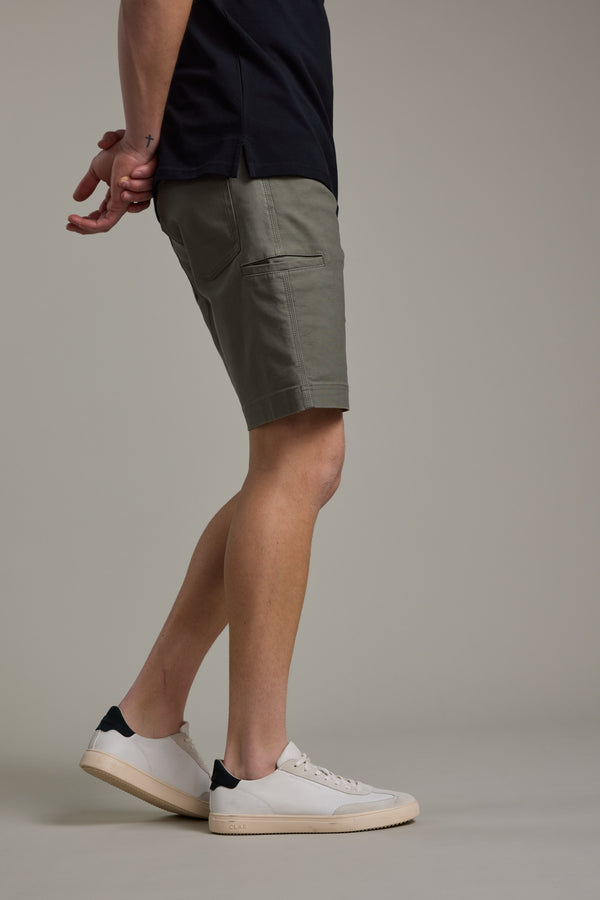 A person wears Barkers Anvil 5Pkt Short in olive green, paired with a black shirt and white sneakers, standing against a plain background with hands clasped behind their back. Only the lower half of their body is visible.