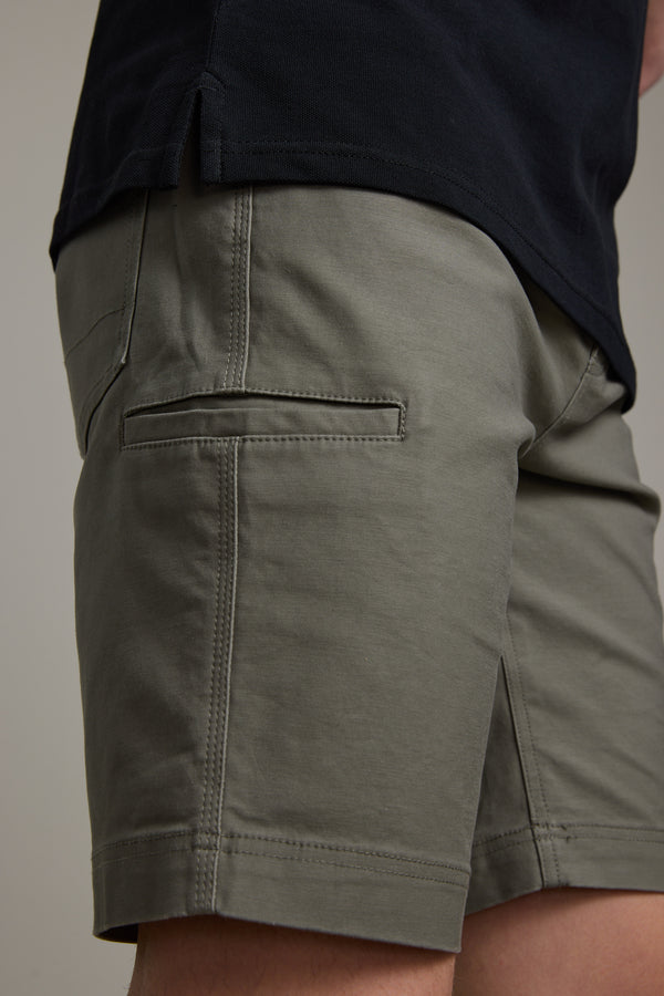 A close-up shows a person wearing olive green Barkers Anvil 5Pkt Shorts with a welt back pocket and elasticated waistband, paired with a black shirt. Only the lower torso and upper legs are visible.