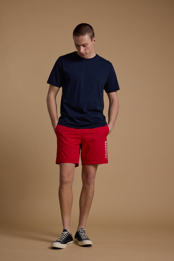 A young man stands against a plain beige background, wearing a navy blue t-shirt and Barkers Weekend Short in red with white lettering—made from recycled polyester for quick dry comfort. He has short hair, looks down, and keeps his hands in his pockets.