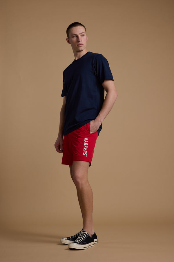 A young man stands against a beige background in a navy t-shirt and Barkers Weekend Short by Barkers, featuring "BARNEYS" vertically on the side, paired with black low-top sneakers. He looks left with one hand in his pocket.