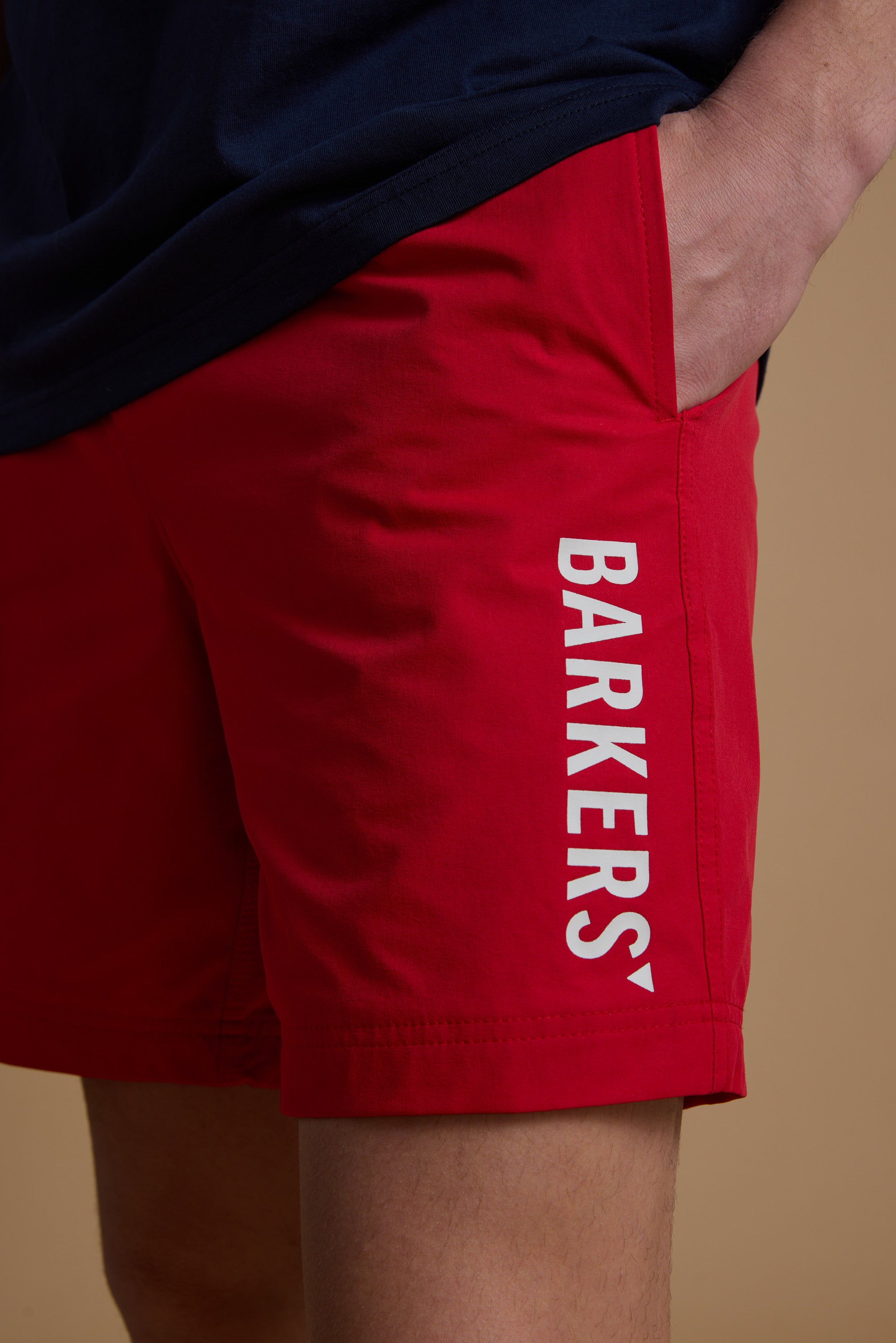 Someone wears bright red Barkers Weekend Shorts—made from recycled polyester, with "BARKERS" in white down the left leg. Their navy shirt is partly visible, and their left hand rests in the pocket of the quick-dry shorts.