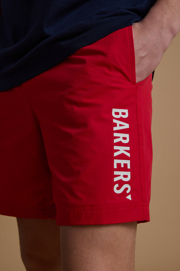 Someone wears bright red Barkers Weekend Shorts—made from recycled polyester, with "BARKERS" in white down the left leg. Their navy shirt is partly visible, and their left hand rests in the pocket of the quick-dry shorts.