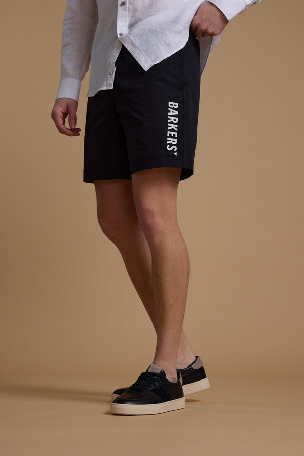 A person in a white button-up shirt, black Barkers Weekend Short by Barkers made from recycled polyester, and black sneakers with white soles stands against a plain beige background.