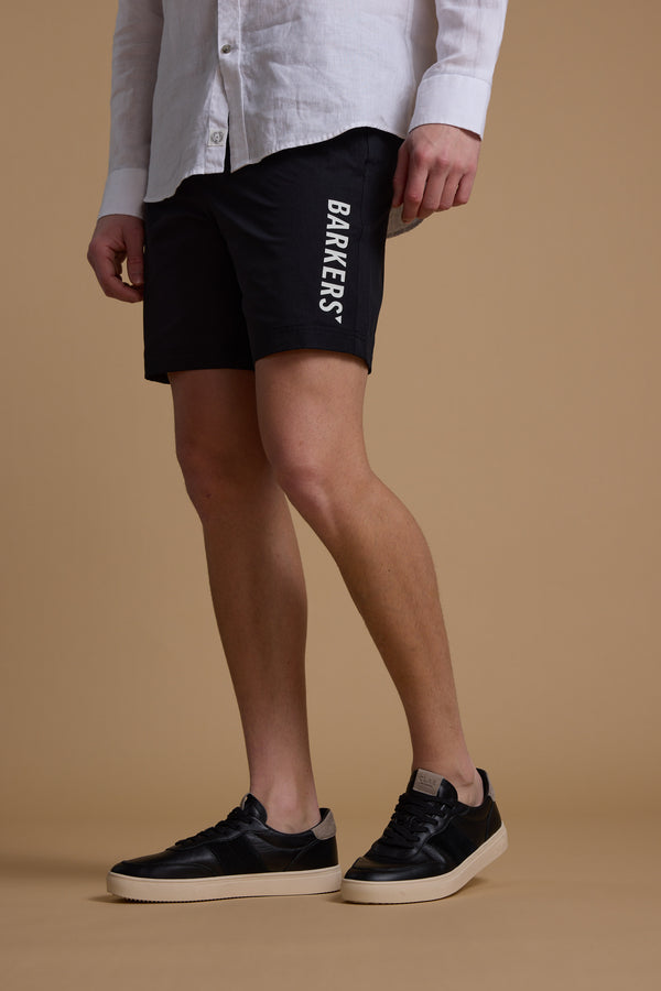 A person in a white button-up shirt, Barkers Weekend Short by Barkers made from recycled polyester, and black sneakers with white soles stands against a plain beige background.