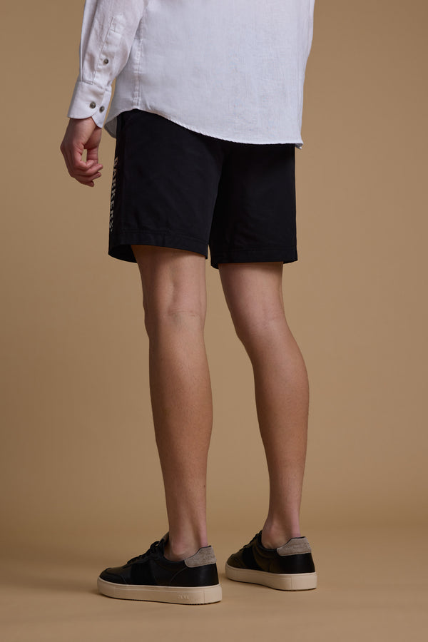 A person stands on a beige surface, seen from behind, wearing a white long-sleeve shirt, Barkers Weekend Short in black recycled polyester, and black low-top sneakers with white soles.