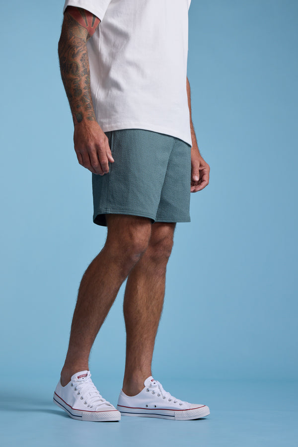 A person stands against a blue background, wearing Barkers' Dehaven Stripe Volley shorts, a white T-shirt, and white sneakers. Only the lower half of their body is visible, with one tattooed arm subtly in view.