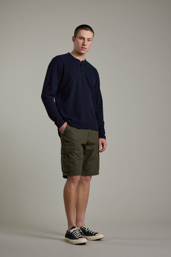 A young man with short hair and a neutral expression stands against a plain background, wearing the Barkers Avenue Waffle Henley in navy, olive cargo shorts, and black low-top sneakers, with his hands in his pockets.