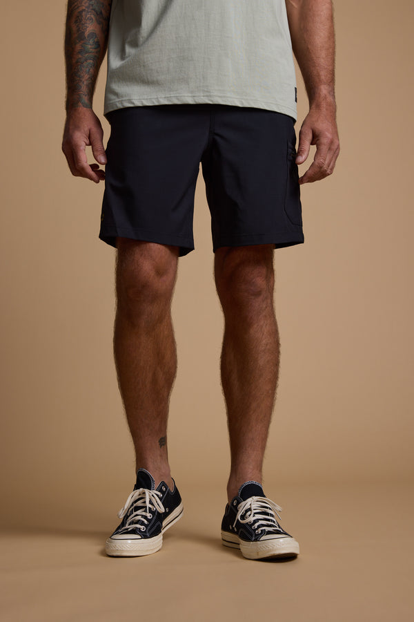 Against a plain beige backdrop, a man with tattooed arms (face not shown) wears a light grey tee and black Legasea Weekend Shorts from Legasea, paired with black low-top sneakers.