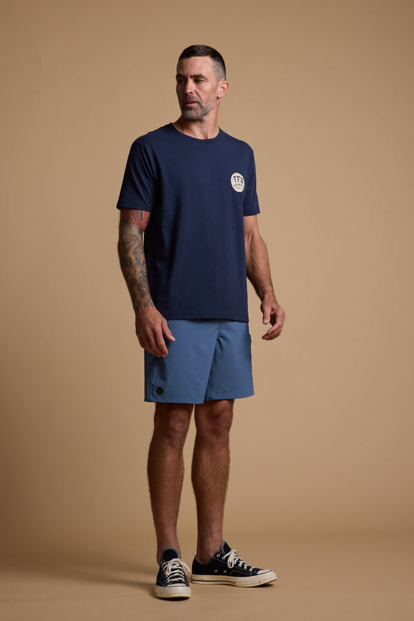 A man stands against a beige background wearing a navy Legasea Snapper Print Tee, blue shorts, and black low-top sneakers. His right arm displays tattoos as he looks slightly to the side with a neutral expression.