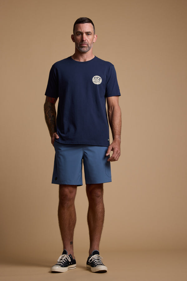 A man stands against a beige background wearing a navy T-shirt, Legasea Weekend Shorts in blue, and black-and-white sneakers. His right hand is in his pocket and he has a neutral expression.
