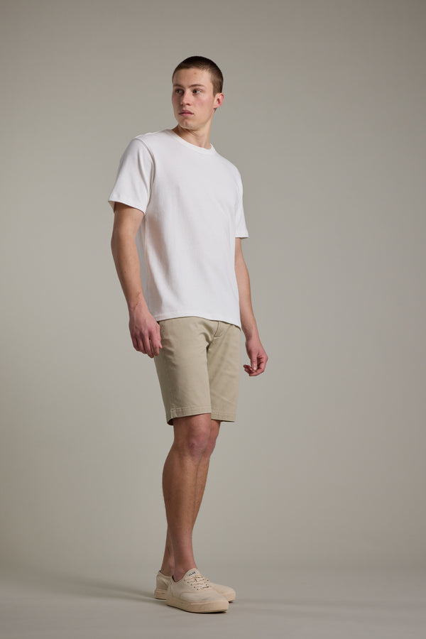 A person stands against a plain background wearing a white short-sleeve t-shirt, Barkers Braddock Walk Short, and beige sneakers, looking slightly to the side.