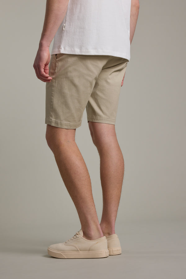 A person stands against a plain background, wearing a white t-shirt and Barkers Braddock Walk Short in beige with matching sneakers. The rear view highlights the relaxed fit and lightweight summer design of these versatile Barkers shorts.