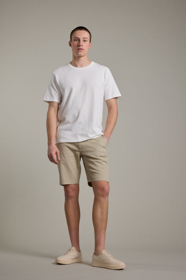 A young man stands against a plain background, wearing Barkers Braddock Walk Shorts in beige with a white T-shirt and matching sneakers—an effortlessly versatile look. He strikes a casual pose, one hand in his pocket and a neutral expression.