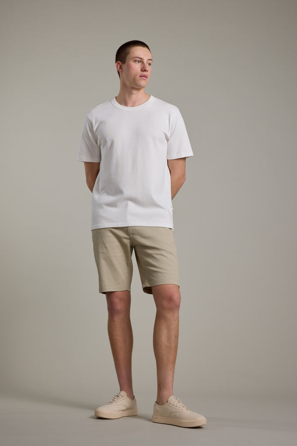 A man stands against a plain background, wearing Barkers Braddock Walk Shorts and a white short-sleeve tee with beige sneakers. His hands are behind his back as he looks slightly to the side.