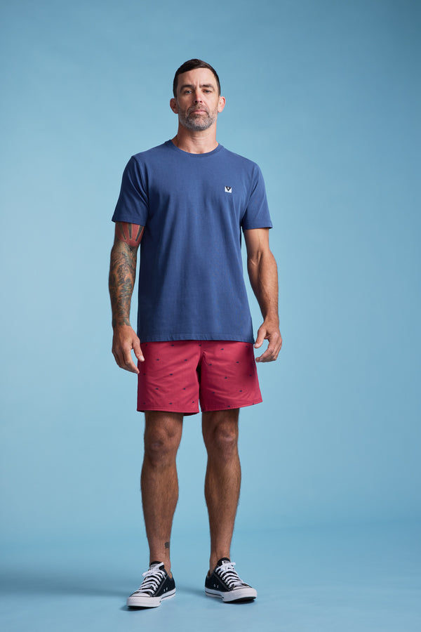 A man with short dark hair and tattooed arms stands against a blue background, wearing Barkers Crayfish Print Volley swim trunks, a blue t-shirt, and black-and-white sneakers.