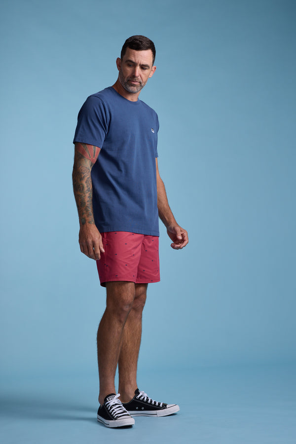 A man with short dark hair and tattoos wears a blue t-shirt, Barkers Crayfish Print Volley shorts made from recycled polyester, and black sneakers while standing against a plain blue background.