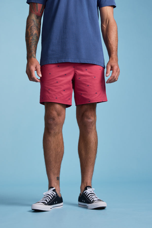 A person stands against a blue background wearing a blue shirt and Barkers Crayfish Print Volley—red quick-dry swim trunks with subtle dark patterns—paired with black low-top sneakers. Tattoos are visible on one forearm.