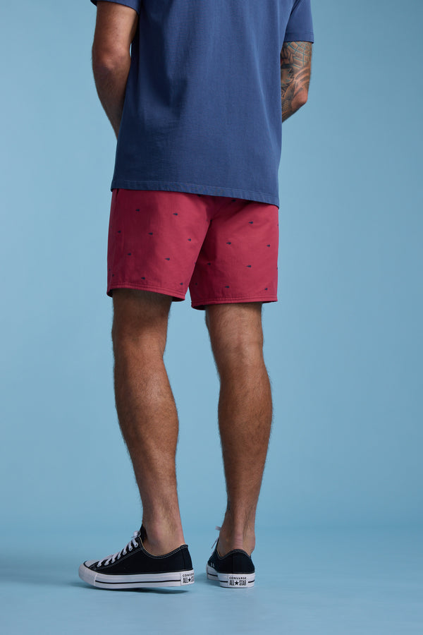 A person stands against a blue background, wearing a blue t-shirt and Barkers Crayfish Print Volley shorts in red with small prints, black low-top sneakers, and has a tattoo on their upper left arm. The casual look is viewed from the back.