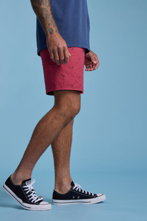 A person with tattooed arms wears a blue shirt, Barkers Crayfish Print Volley shorts in red with small prints, and black Converse sneakers, standing against a plain light blue background.
