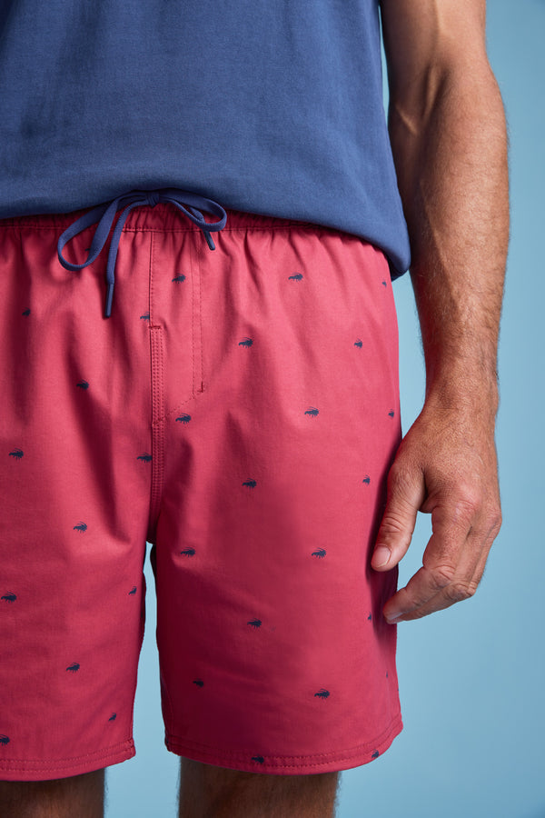 A person wears Barkers’ Crayfish Print Volley—bright pink quick-dry swim trunks with a navy drawstring and small navy crayfish prints—standing against a light blue background. Only the torso and one hand are visible.