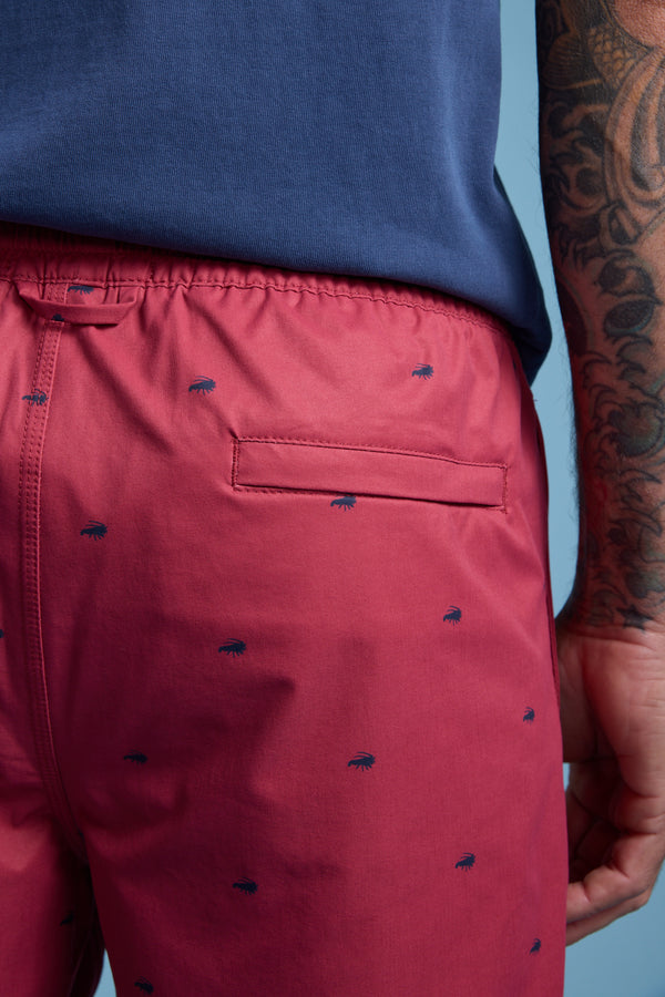A person in Barkers Crayfish Print Volley swim trunks and a blue shirt stands sideways, showing off a tattooed arm and the back pocket of the shorts against a light blue background.
