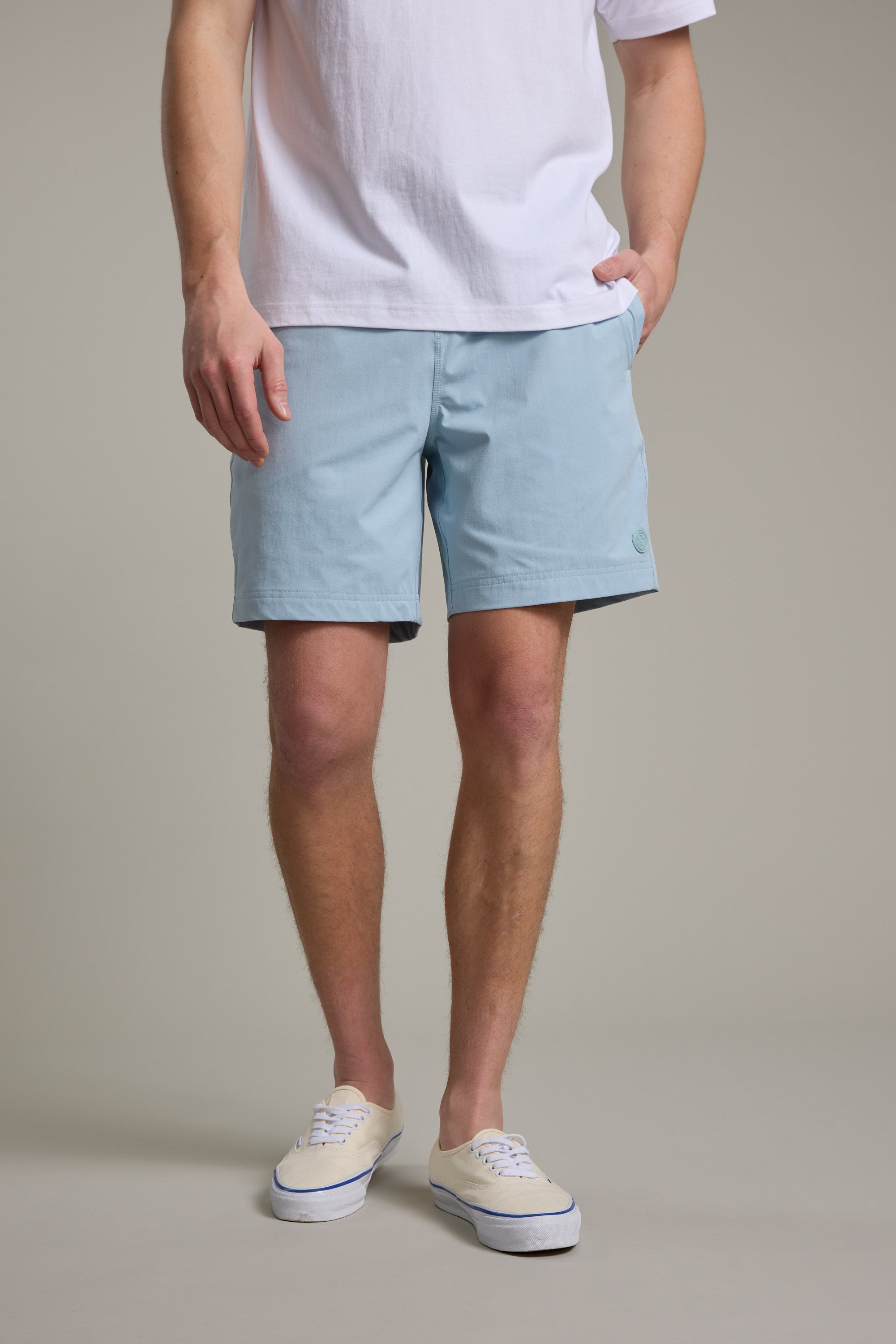 A person models Barkers Davis Weekend Short in light blue recycled polyester with a white T-shirt and sneakers, one hand in pocket, standing against a beige background. Their face is not visible.