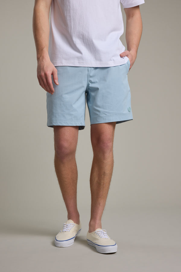 A person models Barkers Davis Weekend Short in light blue recycled polyester with a white T-shirt and sneakers, one hand in pocket, standing against a beige background. Their face is not visible.
