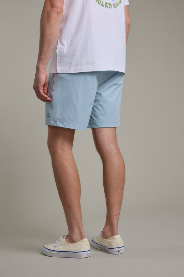 A person stands facing away, wearing a white t-shirt, Davis Weekend Short in light blue by Barkers, and off-white slip-on shoes against a plain neutral background.