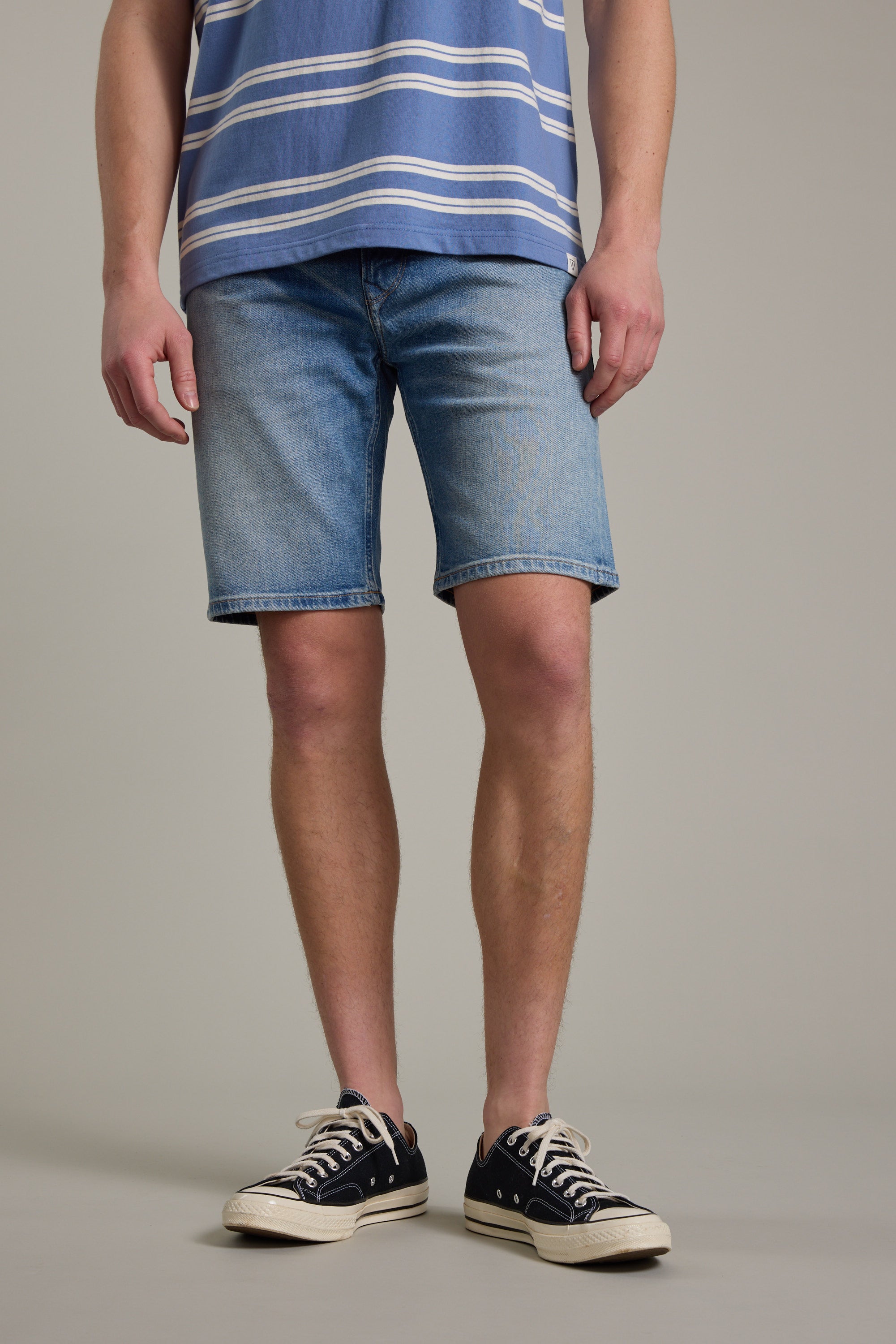 A person wears Barkers' Rover Denim Short—relaxed fit and made from regenerative cotton—with a blue and white striped T-shirt and black low-top sneakers, standing against a plain light background. Their face is not visible.