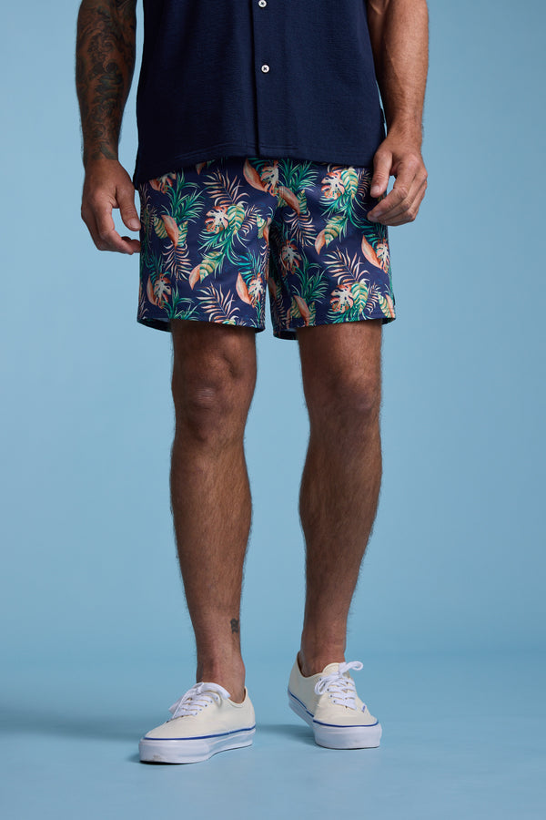 A man, visible from the waist down, stands against a light blue background wearing Barkers Foliage Print Volley shorts made from recycled polyester.