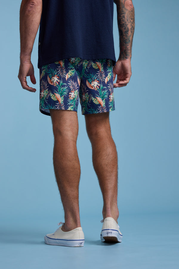 Against a blue background, a person stands with their back to the camera wearing a navy shirt, white sneakers, and Barkers Foliage Print Volley shorts—pineapple and leaf patterned in recycled polyester. A tattoo is visible on one arm.