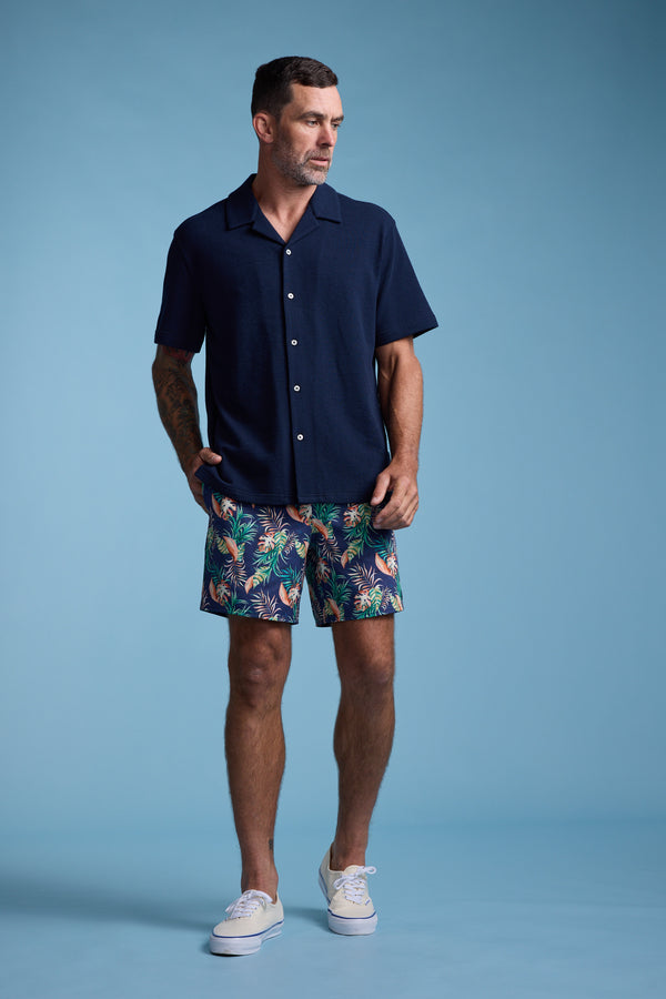 A man with short dark hair and a beard stands against a plain blue background wearing Barkers Foliage Print Volley shorts made from recycled polyester, a navy short-sleeve button-up shirt, and white sneakers, with one hand in his pocket.