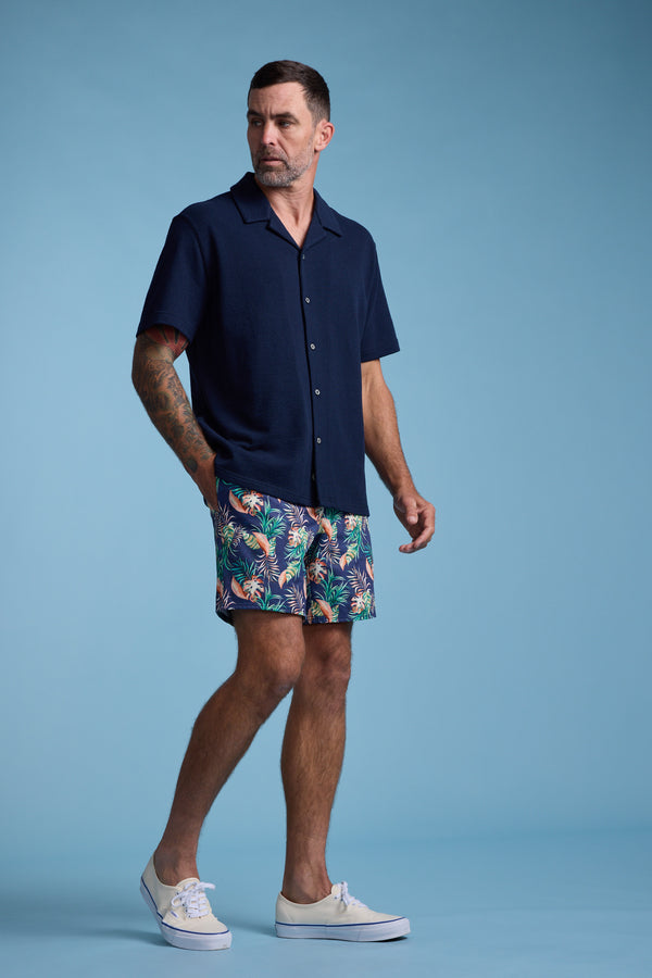 A man with short dark hair, a beard, and arm tattoos stands against a blue background in a navy short-sleeve shirt and Barkers Foliage Print Volley swim trunks with a colorful tropical print, paired with white sneakers.