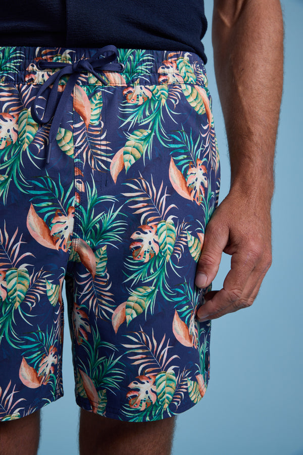 A person wears Barkers Foliage Print Volley shorts—navy blue with a tropical leaf pattern, crafted from recycled polyester—while standing against a light blue background with one hand by their side.