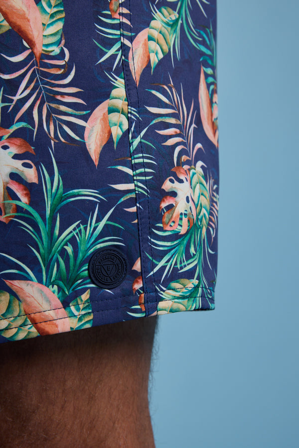 Close-up of a person wearing Barkers Foliage Print Volley shorts, made from recycled polyester with a vibrant green, orange, and white tropical leaf pattern on dark blue, set against a solid light blue background.