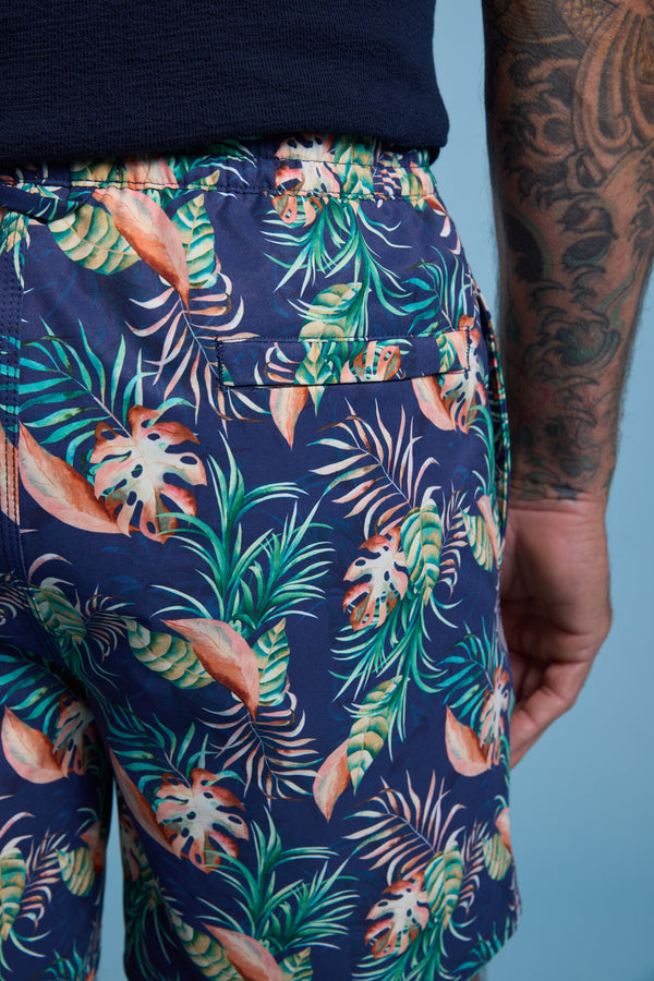 A person in Barkers Foliage Print Volley shorts—navy blue with a colorful tropical leaf print and made from recycled polyester—stands showing a tattoo sleeve against a plain light blue background.