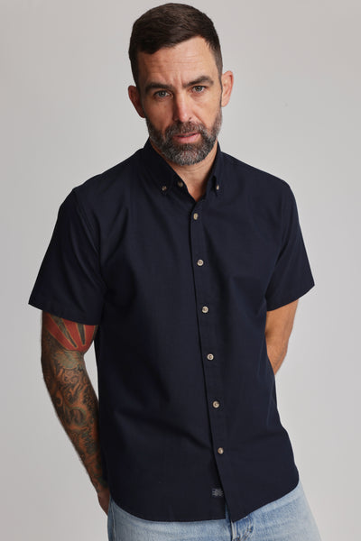 A man with short brown hair and a beard wears the Barkers Archer Oxford Shirt—navy, 100% cotton, button-down collar—with light blue jeans. He stands with one hand in his pocket; tattoos show on his left arm against a plain background.