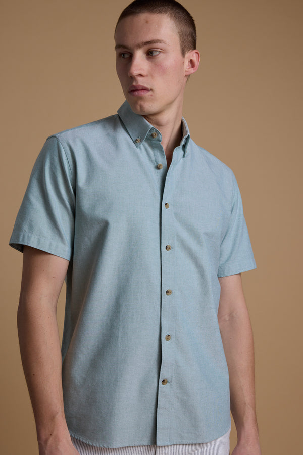 A person models the Barkers Archer Oxford Shirt in light blue—a short-sleeve, button-down collar style—standing against a beige background and looking left with a neutral expression.