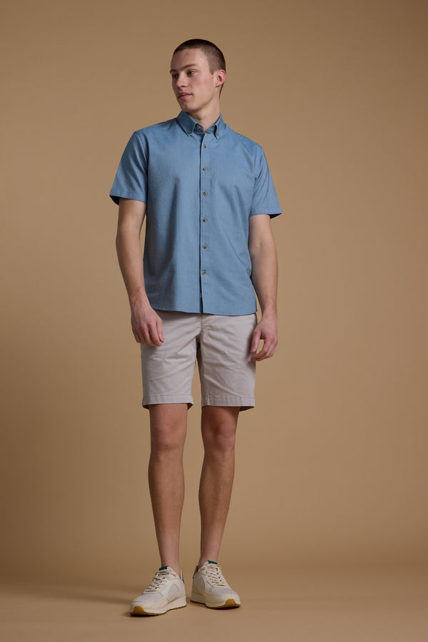 A person stands against a plain tan background, wearing a short-sleeve blue button-up shirt, Barkers Kingly Chino Short in light beige, and cream sneakers. They look slightly to the side with a neutral expression.