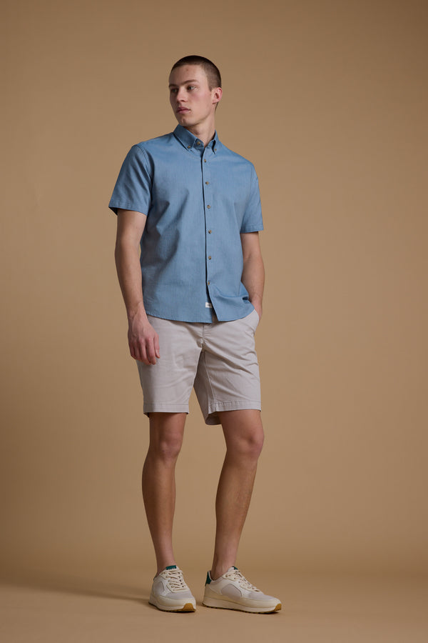 A man stands against a plain beige background wearing the Barkers Archer Oxford Shirt in light blue, paired with light beige shorts and white sneakers. He looks slightly to the side with one hand in his pocket.