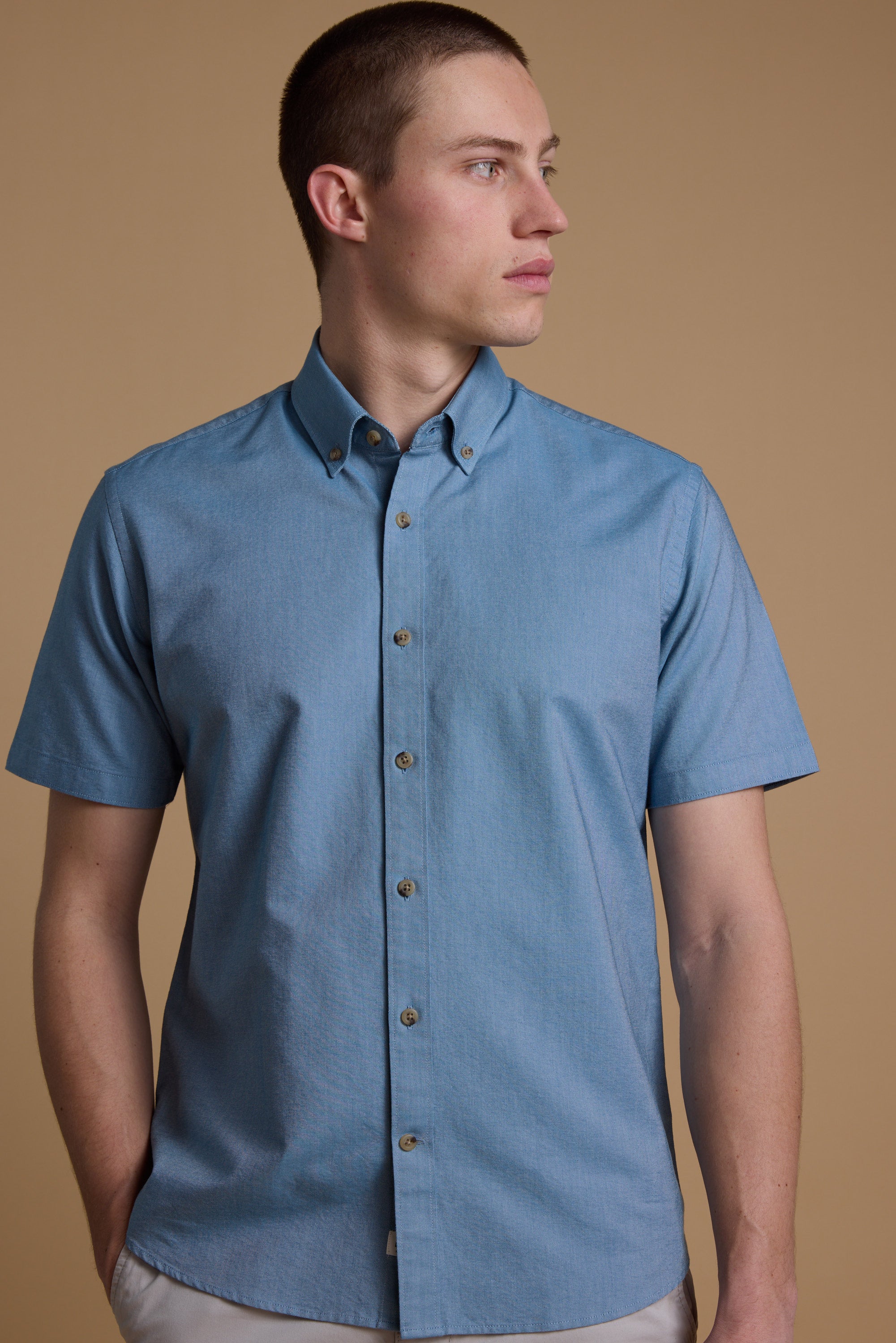 A man stands against a beige background wearing the Barkers Archer Oxford Shirt in light blue, featuring short sleeves and a button-down collar, while looking to his left with a neutral expression.