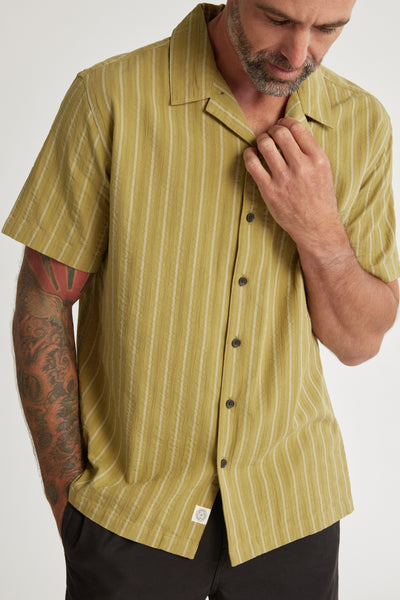 A man with a short beard and a tattooed arm wears the Barkers Goodwin Stripe Cuban Shirt, an olive green, short-sleeved, striped shirt, paired with black pants. He looks down and touches the shirt’s collar with his right hand.