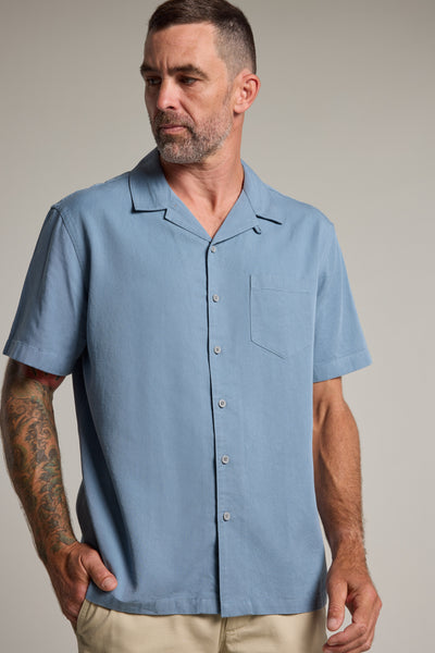 A man with short hair, a beard, and right arm tattoos wears beige pants and the Barkers Perry Cuban Shirt—a light blue Tencel-linen blend men’s summer shirt—standing with one hand in his pocket, looking to his left.