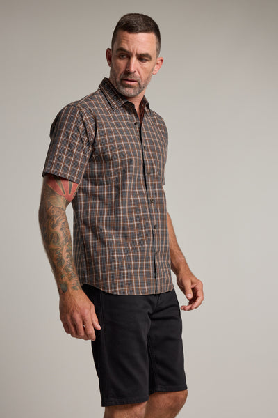 A man with short hair and tattoos on his left arm wears the Barkers Marcus Check Shirt, a classic fit, short-sleeved brown check shirt made from responsibly sourced cotton, paired with black shorts against a plain light gray background.