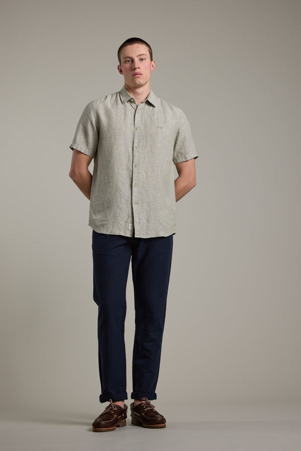 A man stands against a plain background wearing Barkers’ Newman Linen Pant, paired with a light gray short-sleeve button-up shirt and brown shoes. His hands are behind his back and his expression is neutral.